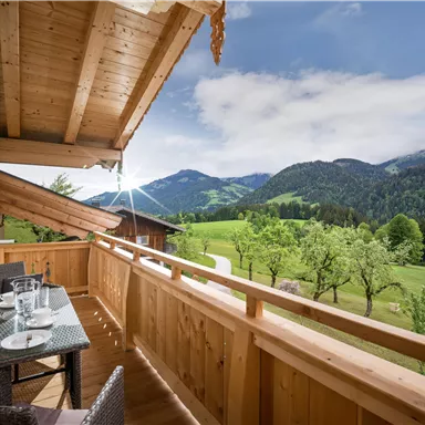 A beautiful balcony with a wooden railing and a table for two. In the background, green meadows and mountains can be seen under a clear sky.