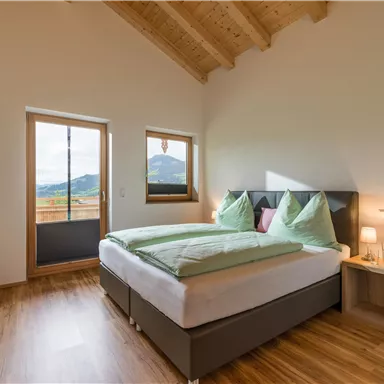 A modern bedroom with a large bed and bright cushions. Through the windows, there is a view of a picturesque landscape.
