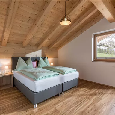 A modern bedroom with a double bed and a wooden ceiling. Large windows offer a view of the outdoors.