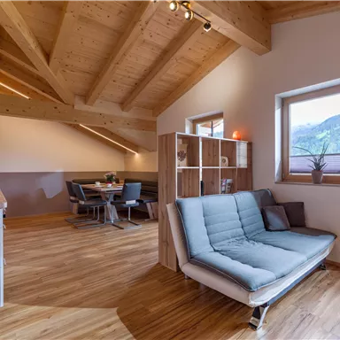 A modern, bright living room with a wooden beam ceiling and a cozy sofa. In the background, there is a dining area with a table and chairs, as well as large windows that offer a view of the mountains.