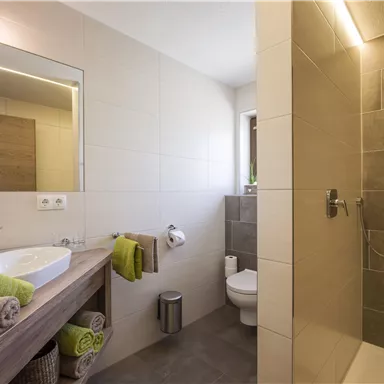 A modern bathroom with a shower and a sink. The walls are tiled in light colors, and there are towels with green accents ready.