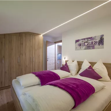 A modern bedroom with two beds and purple pillows. The walls are made of light wood, and there is a pleasant, cozy atmosphere.