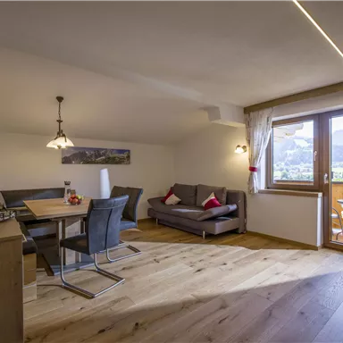 A modern living room with a dining table, a cozy couch, and a large window. The room has warm wooden flooring and offers a view of the mountains.