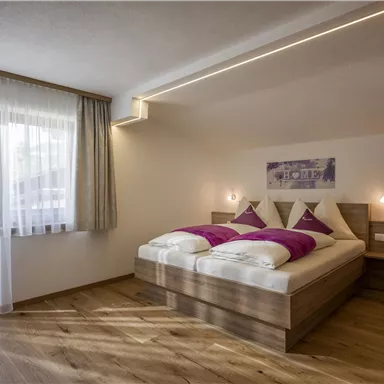 A modern bedroom with a double bed and fresh, purple pillows. The large windows let in a lot of light and create an inviting atmosphere.