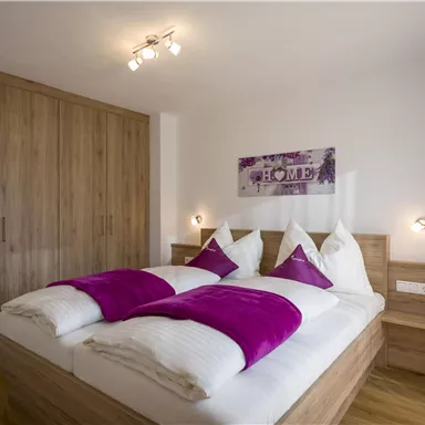 A modern bedroom with a double bed and purple pillows. The room has wooden cabinets and a cozy atmosphere.