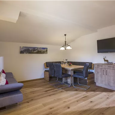 A bright living room with a cozy couch and a dining table for several people. There is a television on the wall, and the floor is made of wood.