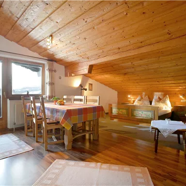 Ein gemütliches Wohnzimmer mit Holzwänden und einem Tisch. Große Fenster lassen viel Licht herein und bieten einen schönen Ausblick.