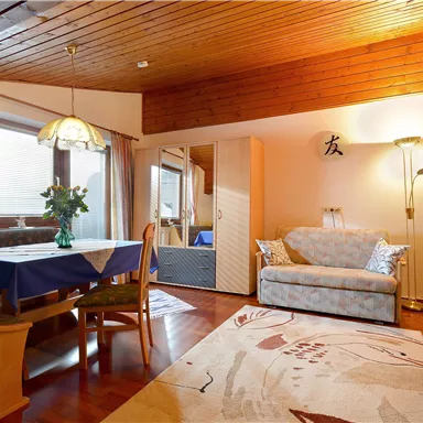 Ein gemütliches Wohnzimmer mit Holzdecke und einem großen Fenster. Es gibt ein Sofa, einen Tisch und einen Fernseher, sowie einen Teppich auf dem Boden.