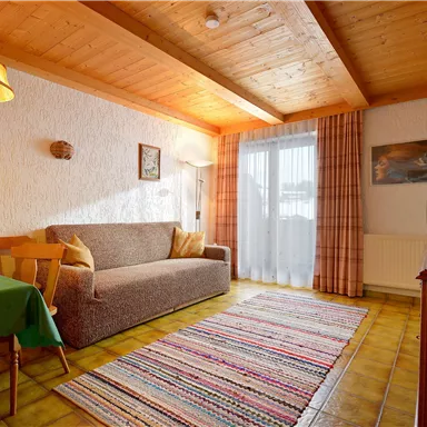 Ein gemütliches Wohnzimmer mit einem Sofa und einem Esstisch. Große Fenster sorgen für viel Tageslicht und eine einladende Atmosphäre.