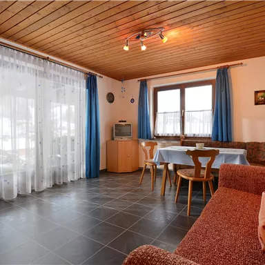 Ein gemütlicher Wohnraum mit Holzdecke, blauen Vorhängen und einer roten Couch. Ein Tisch mit Stühlen steht im Raum, und das Licht ist angenehm warm.