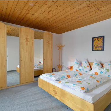 Ein helles Schlafzimmer mit Holzmöbeln und bunten Kissen. Im Hintergrund sind große Spiegelschränke zu sehen.