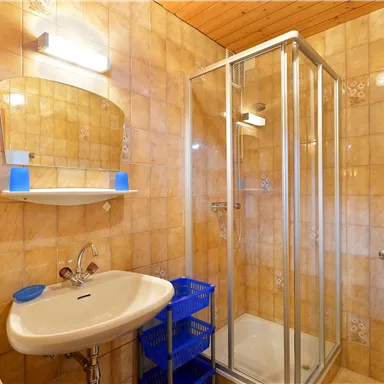 Ein kleines Badezimmer mit einer Glasdusche und einem Waschbecken. Die Wände sind mit hellen Fliesen gefliest und es gibt Handtücher und eine Ablage.