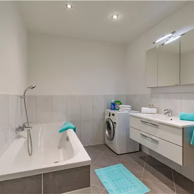 Ein modernes Badezimmer mit einer Badewanne und einem weißen Waschbecken. Es gibt handtücher in Türkistönen und eine Waschmaschine im Raum.