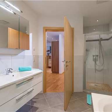 Ein modernes Badezimmer mit einer Dusche und einem Waschbecken. Es gibt helle Farben und Holzdetails, die eine einladende Atmosphäre schaffen.