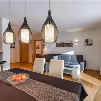 Ein modernes Wohnzimmer mit einem Esstisch und eleganten Lampen. Im Hintergrund ist eine gemütliche Sitzecke zu sehen.