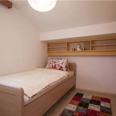 Ein einfaches Schlafzimmer mit einem Einzelbett und einem bunten Teppich. Die Wände sind hell und es gibt Regale über dem Bett.