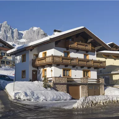 A charming white chalet with wooden railings in a snow-covered landscape. In the background, majestic mountains rise into the clear blue sky.