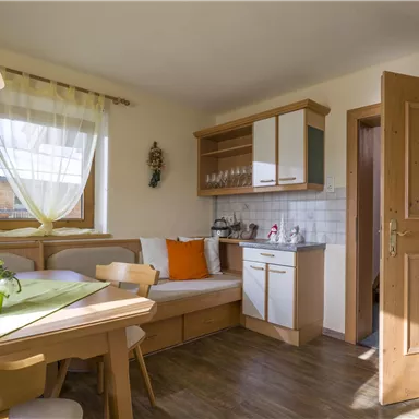 Eine helle Küche mit einem Esstisch und einer gemütlichen Sitzecke. Die Wände sind in warmen Farben gestaltet und die Fenster lassen viel Tageslicht herein.