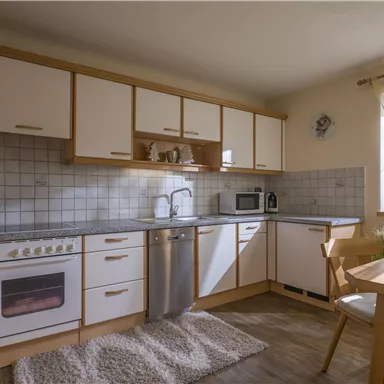 Eine moderne Küche mit weißen Schränken und Holzdetails. Sie ist hell und einladend, mit einem kleinen Esstisch und Fenstern, die viel Licht hereinlassen.