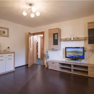 Ein helles Wohnzimmer mit einem Fernseher und modernem Mobiliar. Die Wände sind neutral gehalten und es gibt Holzdetails.