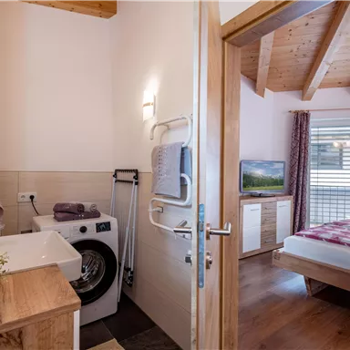A modern bathroom with a washing machine and wooden paneling. In the background, a bedroom with a television and a window is visible.