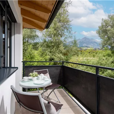 A cozy balcony with a small table and chairs, surrounded by green trees. In the background, gentle mountains and a blue sky can be seen.