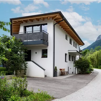 A modern house with a wooden porch and garden, surrounded by trees and mountains. The access road leads to the property.