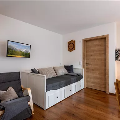 A cozy living space with a sofa bed, a television, and wooden flooring. The walls are bright and inviting, and there is access to another room.