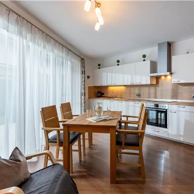A modern kitchen with a bright wooden floor and large windows. There is a dining table with chairs and a calm, inviting atmosphere.
