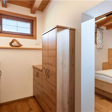 Ein gemütlicher Raum mit Holzschränken und einem Fenster. Im Hintergrund ist ein Schlafzimmer mit einem Bett und sanften Lichtquellen zu sehen.