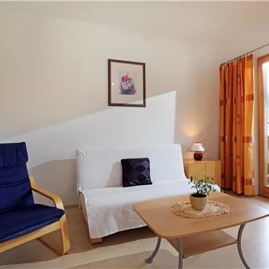 A bright living room with a blue armchair and a white sofa bed.  
Large windows offer a view outside and the room is cozily decorated.