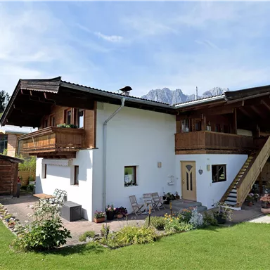 A beautiful two-story house with a balcony and stylish wooden details. Surrounded by green meadows and mountains in the background.