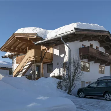 A beautiful chalet in the snow with a car in front of the door. The sky is clear and blue, and the roof is covered with snow.