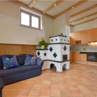 Ein gemütlicher Wohnraum mit einer blauen Couch und einer modernen Küche. Der rustikale Ofen und die Holzdecken verleihen dem Raum eine einladende Atmosphäre.