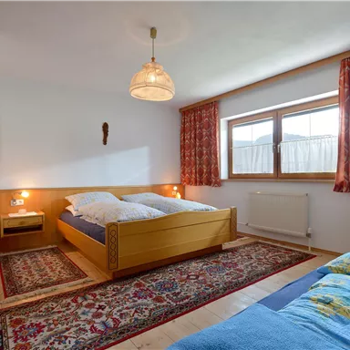 Ein helles Schlafzimmer mit zwei Betten und einem Teppich. Die Fenster sorgen für viel Tageslicht und die Vorhänge sind farbenfroh.