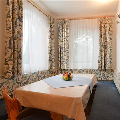 Ein gemütlicher Essbereich mit einem Tisch und stuhlen. Die Fenster sind mit hellen Vorhängen geschmückt, und frisches Obst ist auf dem Tisch platziert.