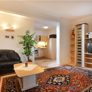 Ein gemütliches Wohnzimmer mit einer schwarzen Couch und gelben Kissen. Im Hintergrund befinden sich ein Esstisch, eine Küche und ein Regal.