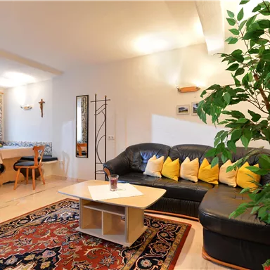 Ein gemütliches Wohnzimmer mit einer schwarzen Couch und gelben Kissen. Ein Tisch und eine grüne Pflanze ergänzen die Einrichtung.