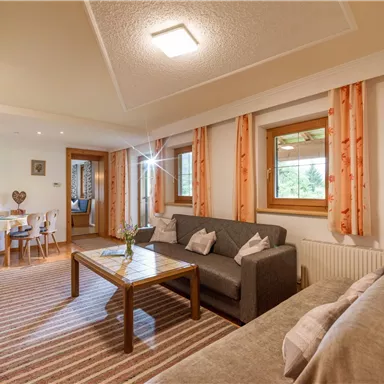 Gemütliches Wohnzimmer mit einem grauen Sofa und einem Holztisch. Große Fenster lassen viel Licht herein und die warmen Farben schaffen eine einladende Atmosphäre.