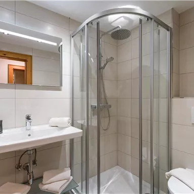 Ein modernes Badezimmer mit einer Dusche aus Glas, einem Waschbecken und einem großen Spiegel. Die Wände sind hell gefliest und es gibt handtücher in der Nähe.