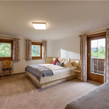 Ein einladendes Schlafzimmer mit einem Doppelbett und einem Einzelbett. Große Fenster sorgen für viel Licht und bieten einen Blick ins Grüne.