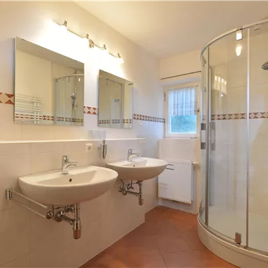 Ein modernes Badezimmer mit zwei Waschbecken und großen Spiegeln. Es gibt eine Dusche in der Ecke und helle, neutrale Farben an den Wänden.