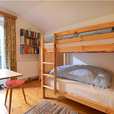 Ein gemütliches Schlafzimmer mit einem Etagenbett und einem kleinen Tisch. Der Raum hat große Fenster und einen Bücherregal.