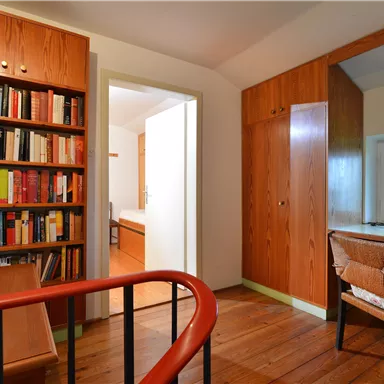 Ein gemütliches Arbeitszimmer mit einem Bücherregal und einem Schreibtisch. Das Zimmer hat holzverkleidete Wände und ein Fenster mit Blick nach draußen.