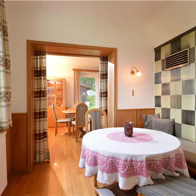 Ein gemütlicher Raum mit einem runden Tisch und einer eleganten Tischdecke. Im Hintergrund sind Stühle und eine dekorative Wand sichtbar.