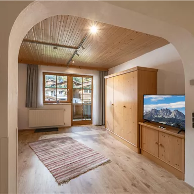 A cozy living space with a wooden ceiling and a large window. A TV and a wooden cabinet can also be seen in the room.