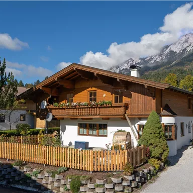 A traditional Austrian house with a wooden balcony and beautiful floral decoration. In the background, majestic mountains can be seen under a blue sky.