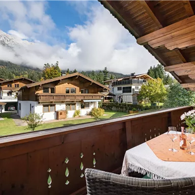 A cozy balcony with a table and two glasses, surrounded by a picturesque mountain landscape. In the background, traditional wooden houses and lush greenery can be seen.
