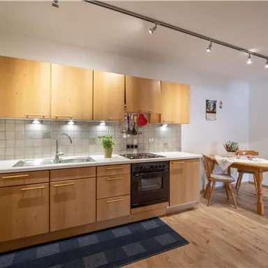 A modern kitchen with wooden furniture and a white countertop. There is a dining table and a carpet on the floor.