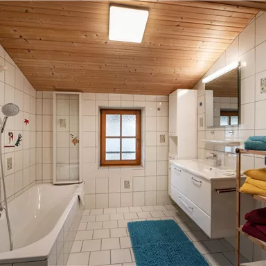 A modern bathroom with a bathtub and a shower. There are bright tiles, a wooden ceiling, and colorful towels.